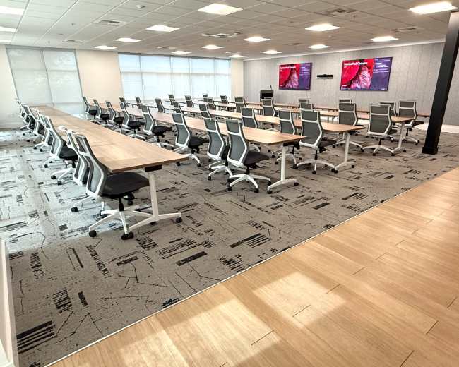 The image shows a modern conference room with rows of tables and chairs, equipped with two wall-mounted screens.