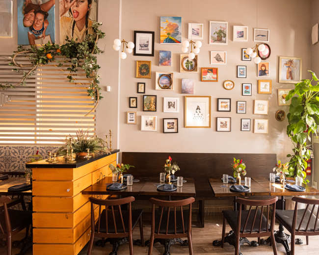 A restaurant interior features a wooden table with chairs arranged in front of a wall adorned with various framed artworks and photographs, while greenery decorates a nearby corner.