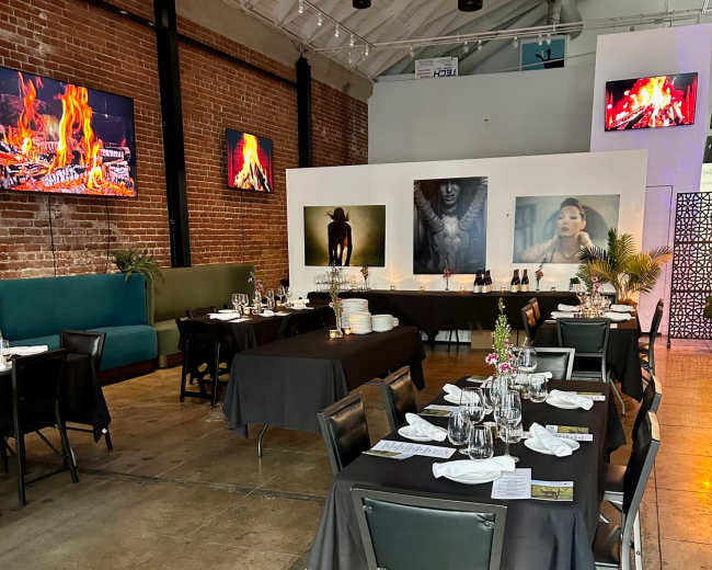The image shows an indoor restaurant space with dark tables set for dining, a brick wall, and large screens displaying fireplace visuals.
