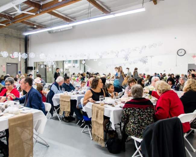 A large group of people sit at tables in a spacious hall, enjoying a meal together during a gathering.