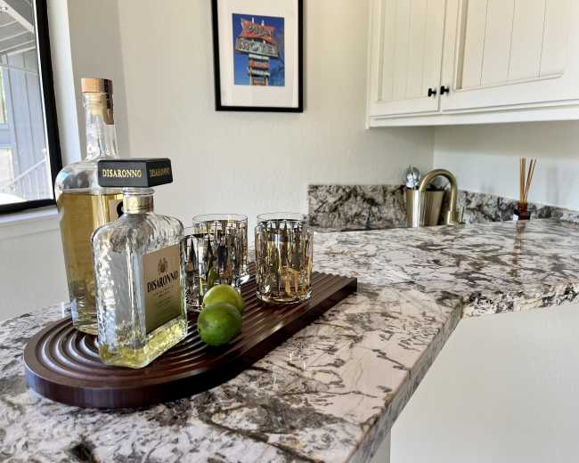 A wooden tray holds a bottle of Disaronno, several glasses, and a lime on a polished marble countertop in a kitchen.