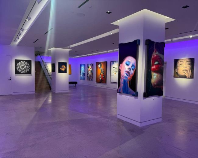 The image shows an art gallery with illuminated walls displaying various framed artworks and a staircase in the background.