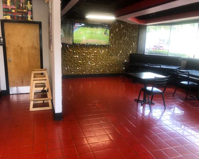The image shows an empty dining area of a restaurant with red tiled flooring, a television displaying a sports game, and a few scattered chairs and tables.