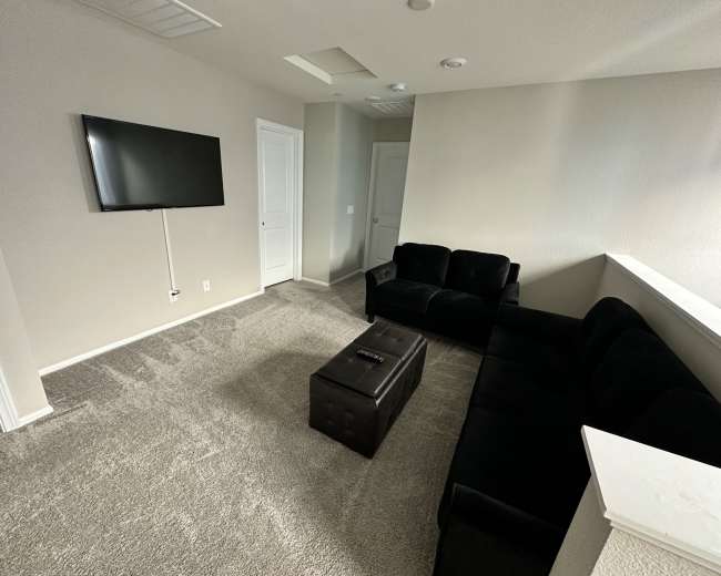The image shows a small living room with a black couch, a coffee table, a television mounted on the wall, and light-colored walls and carpet.