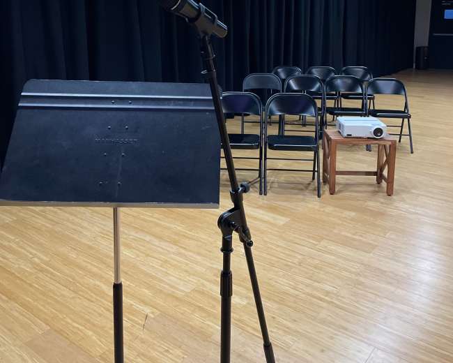 A microphone is mounted on a stand in front of a music stand, with a row of empty chairs and a projector on a small table in the background.