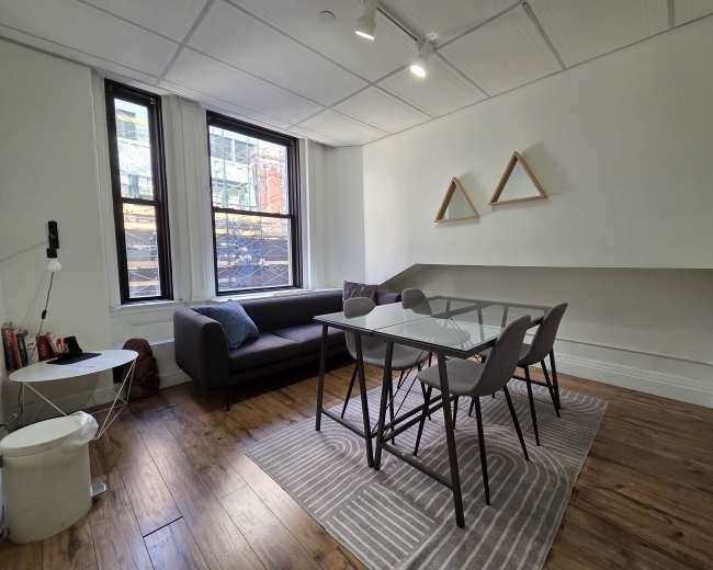 The image shows a modern, minimalist workspace featuring a sofa, a glass dining table with four chairs, and triangular wall decorations, with natural light coming in through a large window.