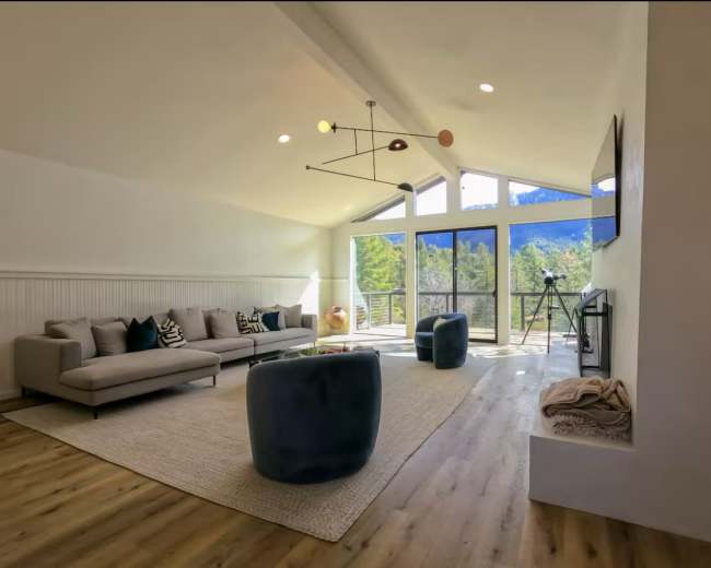 The image shows a modern living room with a light-colored sofa, two dark blue ottomans, and large windows that overlook a mountain view.