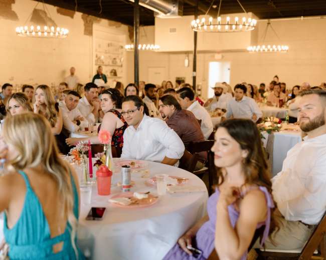 A group of guests sit at elegantly dressed tables during a gathering in a well-lit venue.