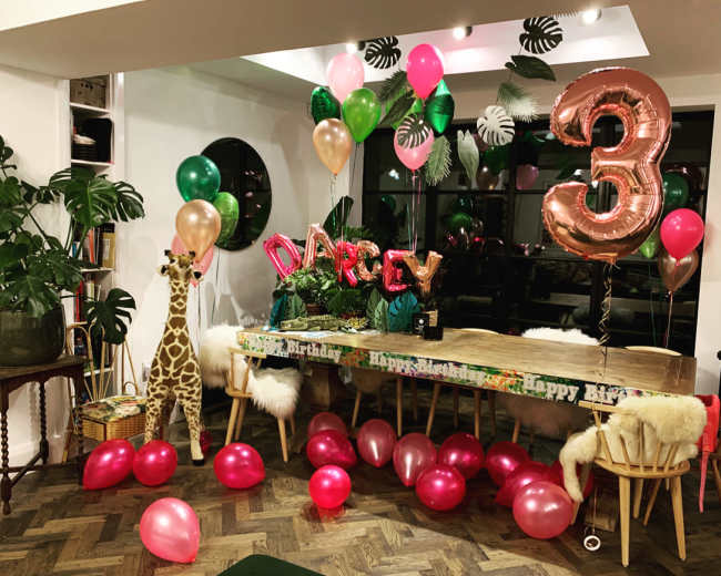 A birthday celebration scene features a table decorated with balloons and themed decorations, along with a stuffed giraffe and scattered pink balloons on the floor.