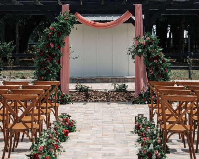 The image shows a wedding ceremony setup with wooden chairs arranged in rows facing a decorated archway draped in fabric and surrounded by floral arrangements.