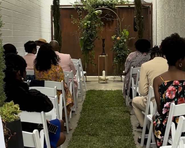 A wedding ceremony setup features white chairs arranged in rows along a grassy aisle leading to a decorated archway.