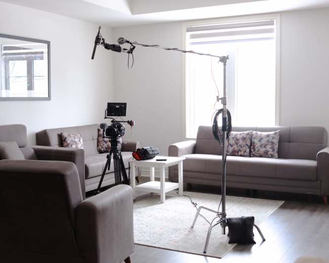 A living room setup for filming, featuring two gray sofas, a coffee table, and camera equipment on a tripod.