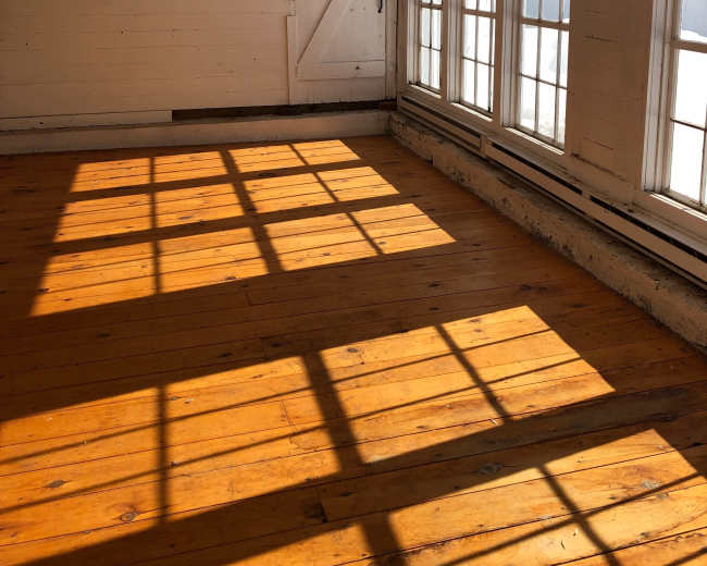 The image shows an empty room with wooden floors and large windows casting shadows on the walls.