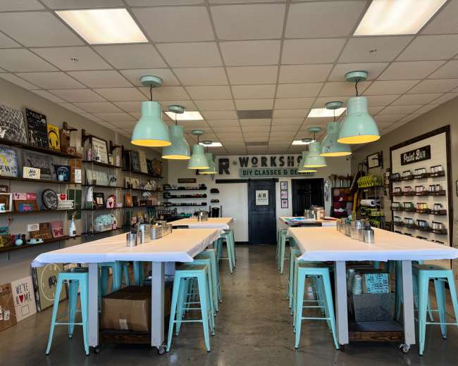 The image shows a brightly lit workshop space with multiple work tables, teal stools, and shelves lined with art supplies and decor items.