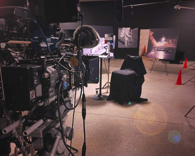 A camera setup with microphones points toward a covered chair and photographs displayed on easels in a dimly lit studio.