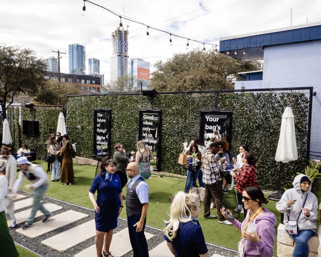 A group of people socialize and mingle in a lively outdoor space adorned with greenery and string lights, while city buildings loom in the background.