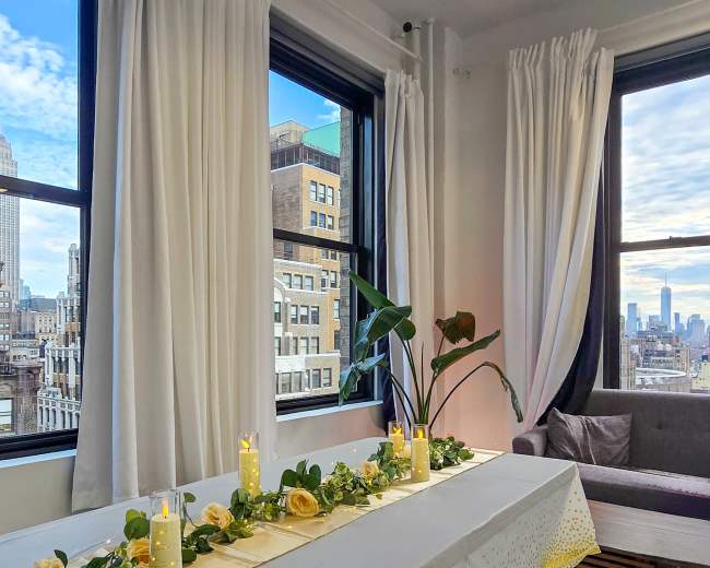 The scene features a table set with candles and greenery near large windows that overlook a cityscape, including the Empire State Building.