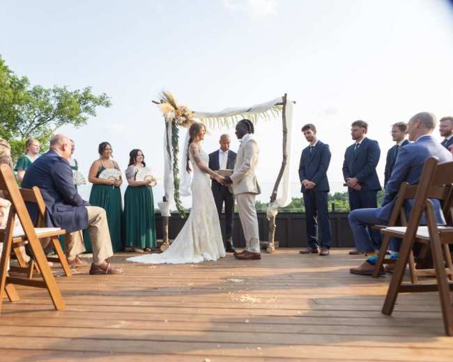 A bride and groom stand under an arch while exchanging vows during an outdoor wedding ceremony, surrounded by seated guests.