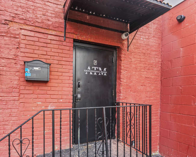 The image shows a black door with a mailbox, set against a red brick wall, with a metal railing leading up to the door.