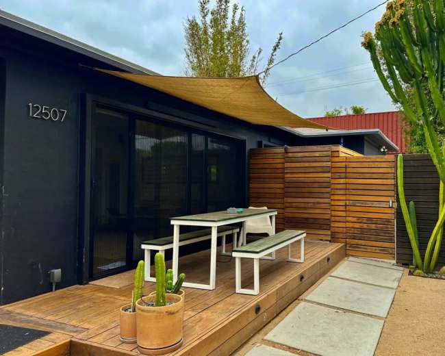 The image shows a modern outdoor patio area with wooden decking, a shaded table and benches, and tall green cacti.