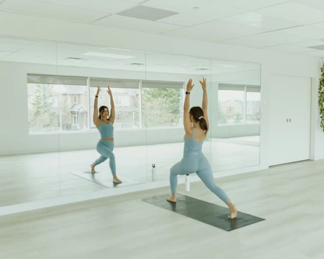 A person practices yoga in a mirrored studio with large windows and green plants on the wall.