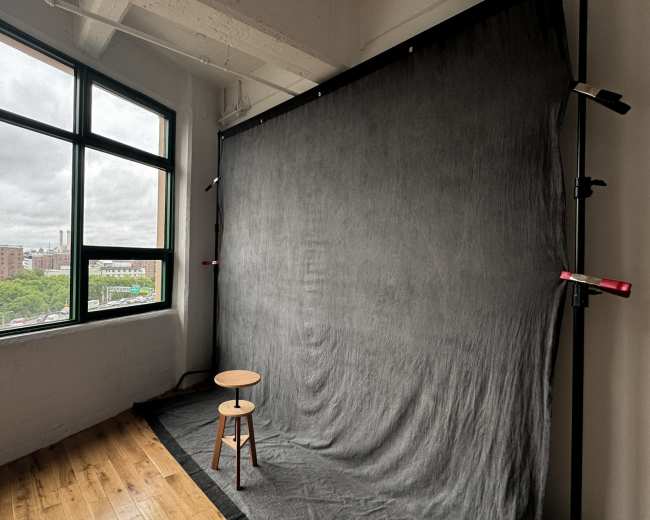 The image shows a photography studio with a black backdrop, a wooden stool, and a large window revealing an urban landscape outside.