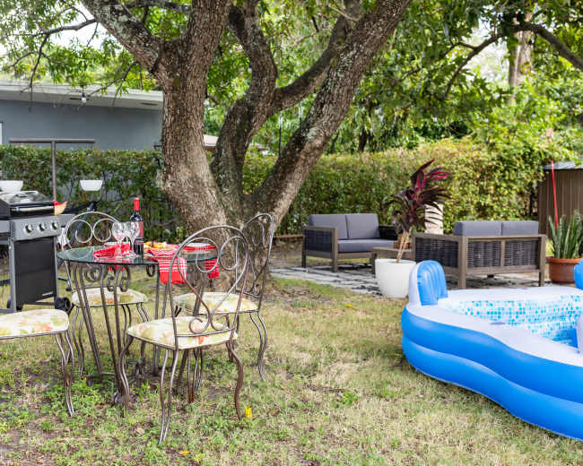 A backyard setup features a grill, a table with chairs, a small inflatable pool, and lounge seating beneath a tree.