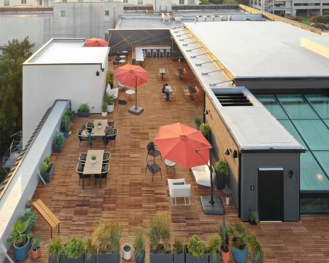 A spacious rooftop terrace with wooden flooring, outdoor furniture, and several red umbrellas, overlooking a cityscape at sunrise.