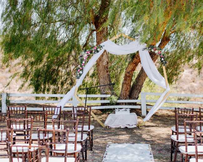 A wedding ceremony setup features an elegant altar draped in white fabric under two large trees, with rows of chairs arranged in front.