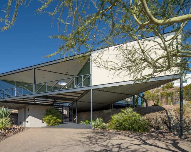 The image shows a modern, elevated house with a distinct angular design, supported by pillars, surrounded by desert landscaping.