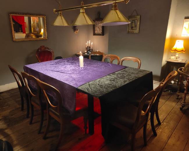 A large table covered with a dark purple cloth sits in a dimly lit room surrounded by chairs, with a candle in the center and warm lighting from a lamp in the corner.