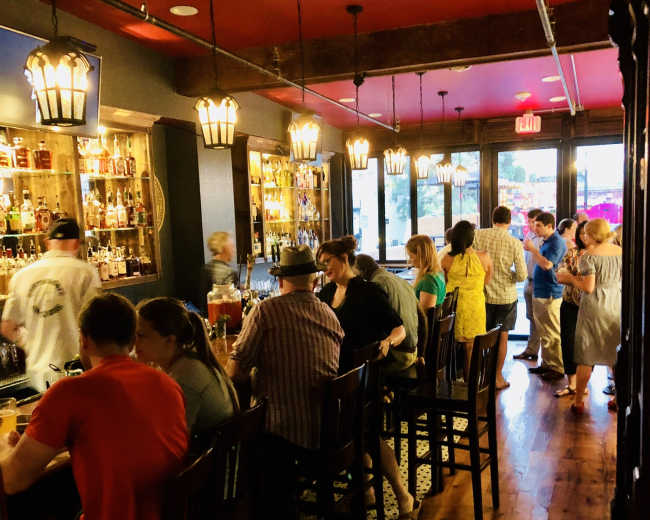 The image shows a busy bar interior with patrons seated at a long counter and others standing near shelves filled with bottles.
