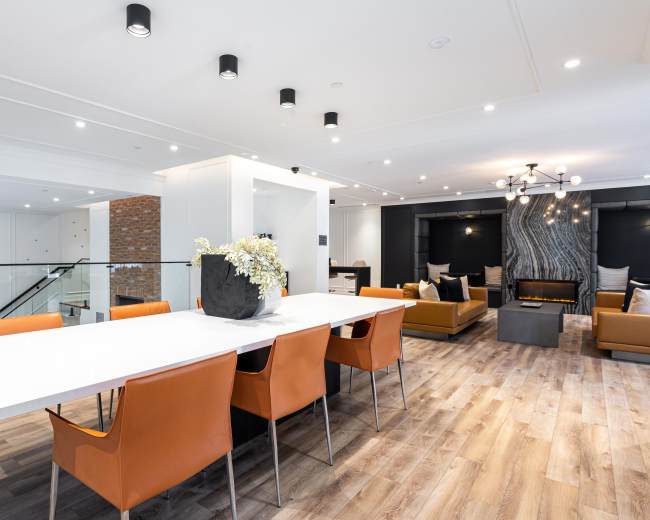 A modern dining area features a long white table surrounded by orange chairs, with a living space and fireplace visible in the background.