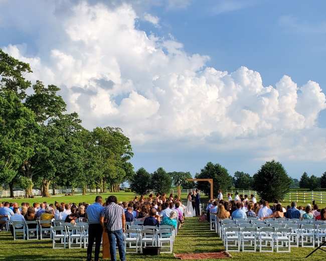 A wedding ceremony is taking place outdoors with white chairs arranged in rows facing an arch, under a bright sky filled with clouds.