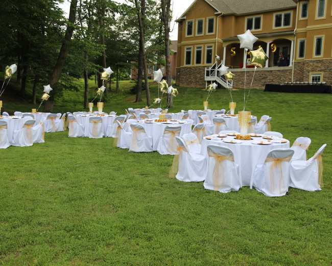 A set of tables covered with white linens and adorned with centerpieces and balloons is arranged on a grassy area near a large house.