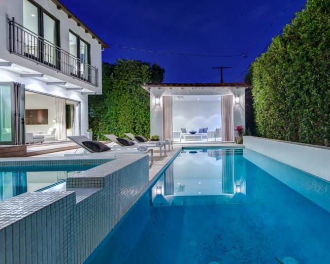 A modern home features an illuminated pool surrounded by lounge chairs, with lush greenery and a stylish outdoor seating area visible in the background.