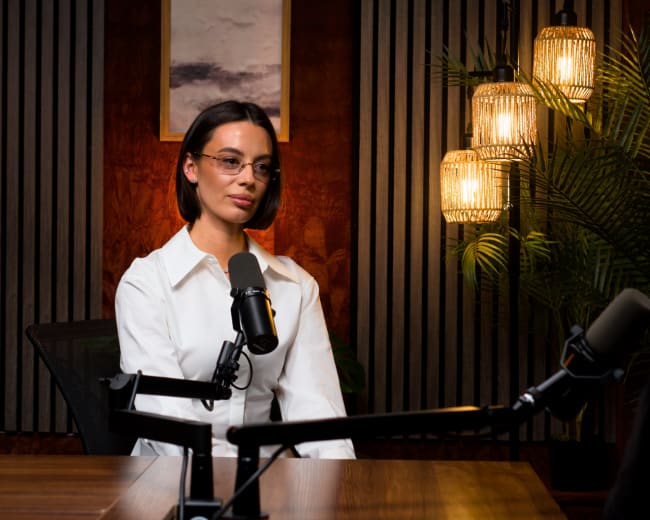 A woman wearing a white shirt sits at a desk with microphones in a well-lit studio featuring wood paneling and hanging lights.