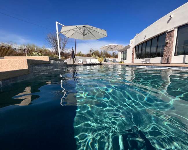 The image shows a clear swimming pool with reflections of umbrellas and a house against a blue sky.