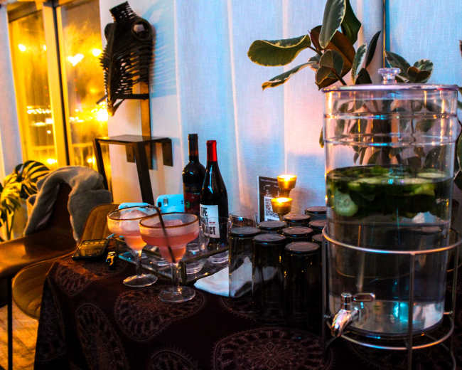 A table is set with a drink dispenser filled with water and cucumbers, alongside bottles of wine, glasses, and candles in a dimly lit room.
