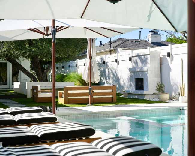 A crystal-clear swimming pool reflects the sun, surrounded by lounge chairs and a landscaped yard with a modern seating area and an outdoor fireplace.