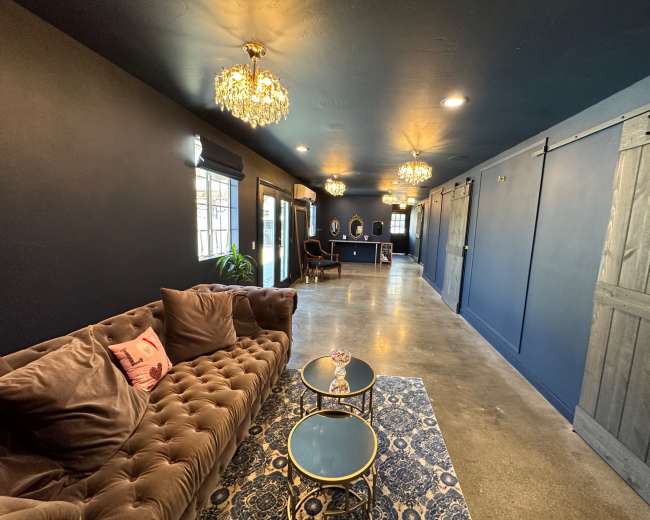 A long, narrow lounge area featuring a tufted brown sofa, two small coffee tables, and a dark blue wall with elegant chandeliers and sliding barn doors.
