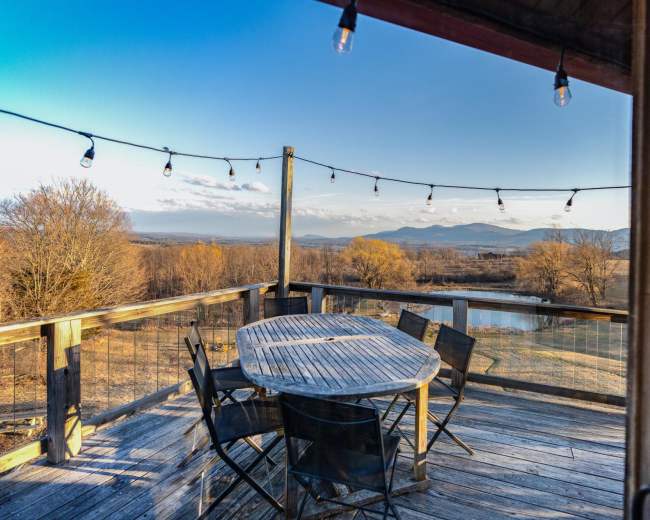 A wooden deck with a circular table and black chairs overlooks a landscape of mountains and a pond, illuminated by string lights.
