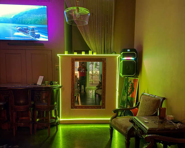 The image shows a cozy bar area featuring a brightly lit mirror with neon green accents, a television displaying a scenic view, and elegant seating arrangements.