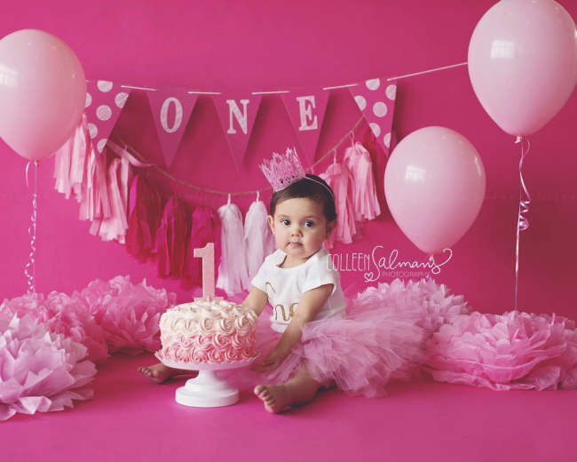 A baby girl sits in front of a pink backdrop adorned with balloons, a decorative banner, and a cake topped with the number one.