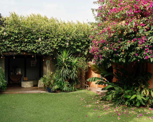A lush garden features vibrant plants and flowers, surrounded by a green lawn and a shaded seating area with string lights.
