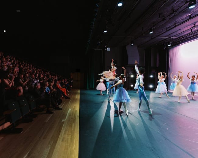 A ballet performance takes place on stage while an audience watches from a seated area.
