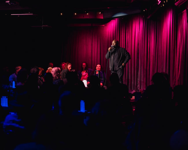 A comedian stands on stage in front of an audience at a dimly lit comedy club with pink and purple lighting.