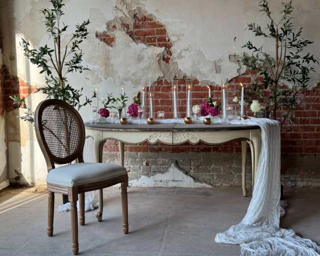 A simple wooden table is adorned with flowers and candles, accompanied by a single chair, against a backdrop of exposed brick walls.