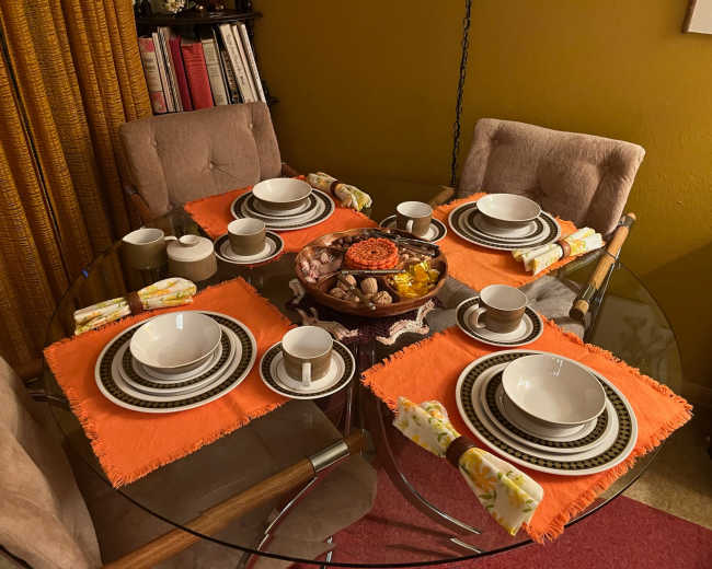 A circular glass dining table set for a meal with four place settings on orange placemats, featuring white bowls, cups, and a round platter of food in the center.