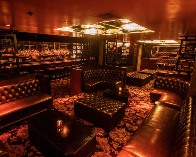 The image shows a dimly lit lounge with dark leather seating, a bar area, and an ornate ceiling fixture.